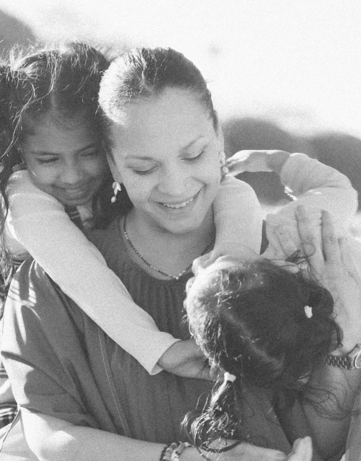 Black and white photo of a mother joyfully embracing her two daughters in soft sunlight