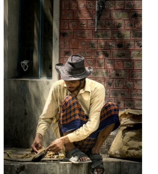 Documentary portrait of a man in a hat preparing food beside a brick wall