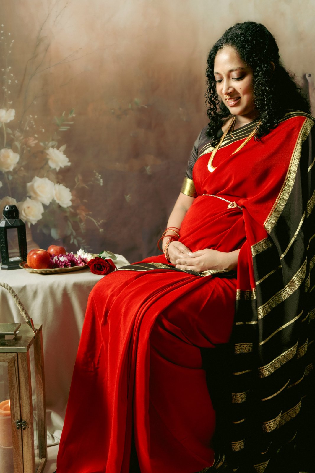 Expectant mother in a red and gold saree cradling her baby bump in a warm, candlelit maternity session